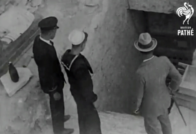 A black and white screenshot showing three men looking into a dark tomb