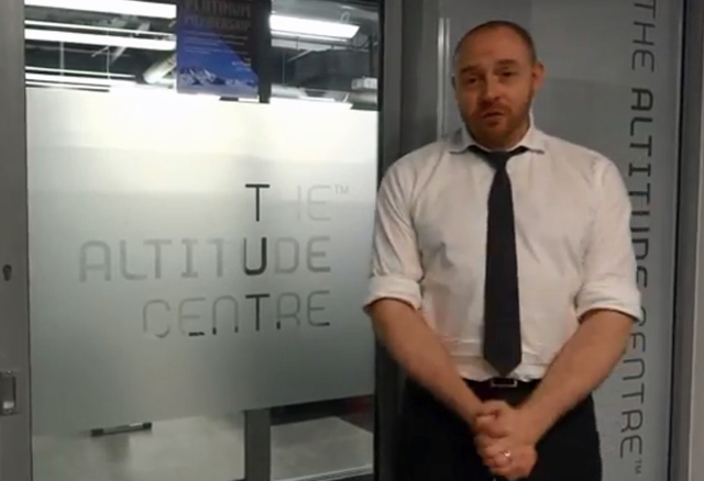 A screenshot showing a man standing in a grey room.