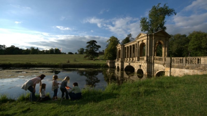 Children and their mums are standing on the edge of the Octagon Lake, using a sweep net to catch any insects as part of a nature activity. Behind them is a large bridge that crosses the river in a garden.