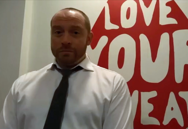 A screenshot showing a man in front of a white wall with some writing on it. 
