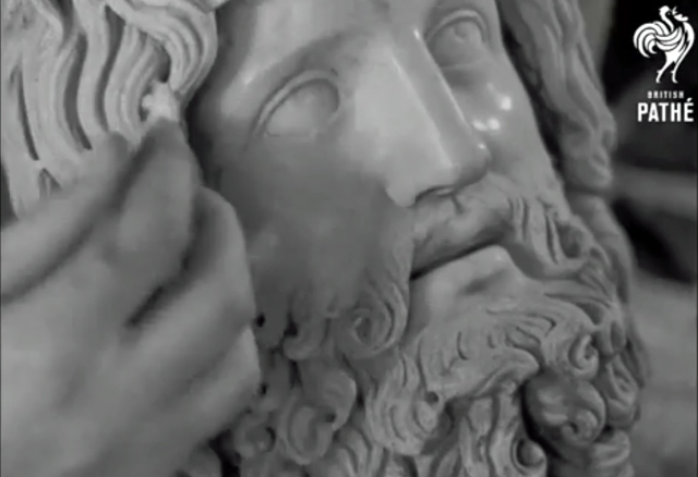 A black and white screenshot showing a curator handling a stone head