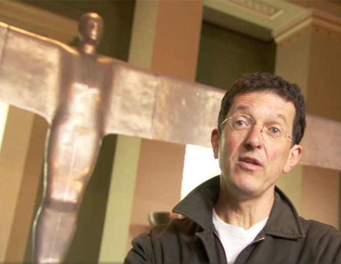 A photograph showing a man with glasses in front of a model of the Angel of the North. 