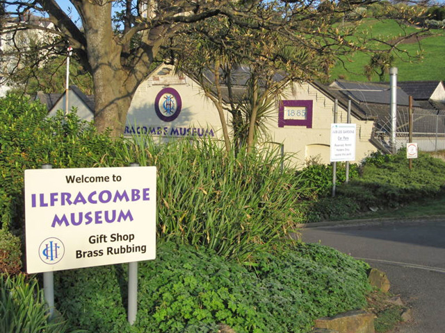 View of a cream coloured building partly obscured by greenery and a sign reading Ilfracombe Museum